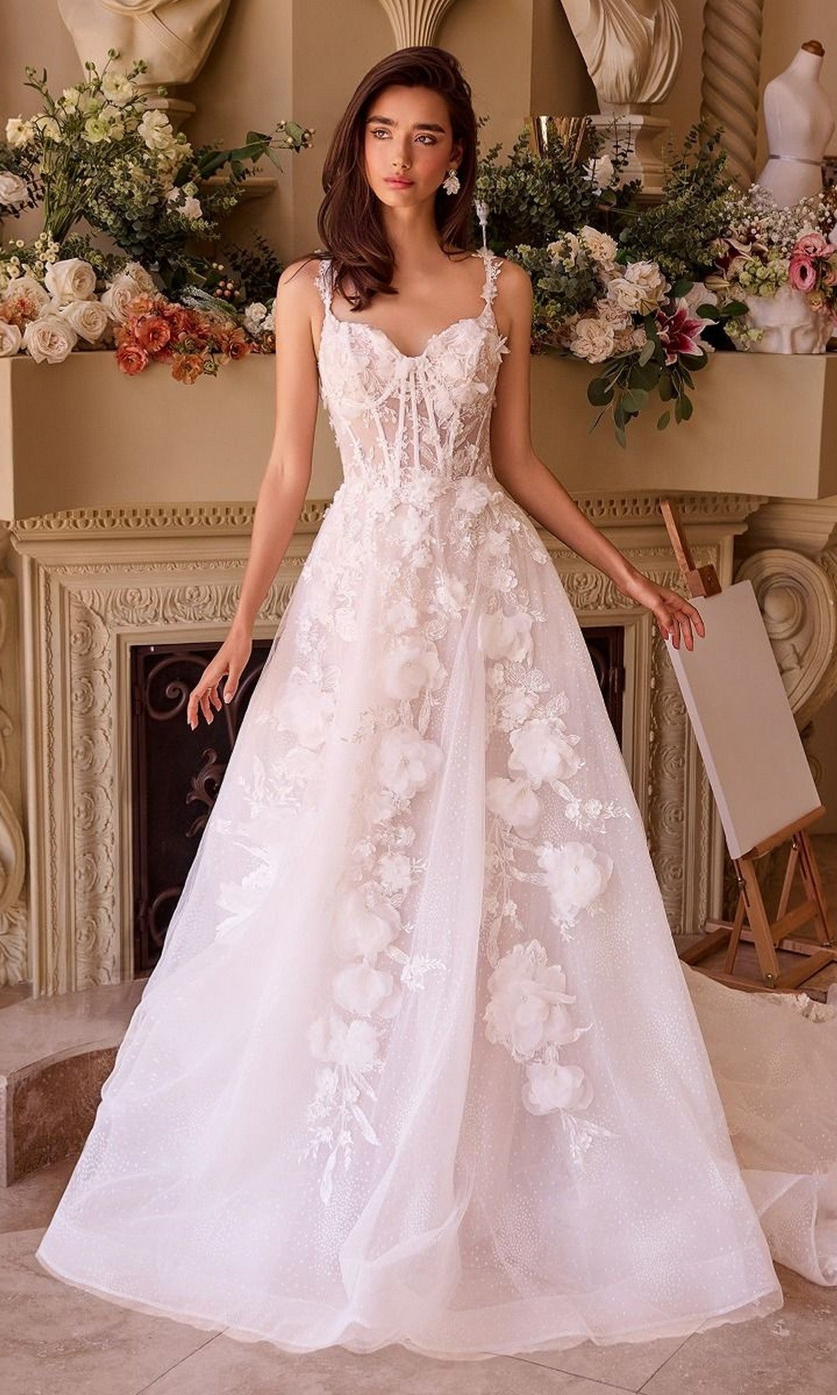 A woman stands before a decorated mantel, wearing the Andrea and Leo WL045 long wedding dress featuring a white lace corset bodice and flowing tulle A-line skirt with delicate floral embellishments.
